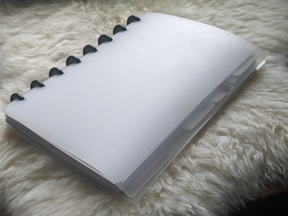 Black plastic discs intersect with a stack of paper, holding translucent plastic cover sheets as the covers of the book. The covers overhang the edge of the paper to protect some tabbed dividers. The whole is presented on a sheepskin.